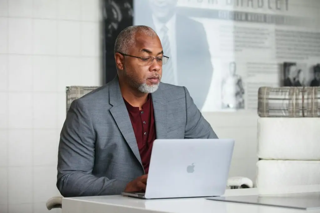 A senior adult man with a gray beard working remotely on a laptop indoors wearing a blazer.